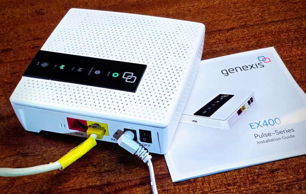 Ein Wlan-Router steht auf einem Holztisch, darunter eein Anleitungs-Büchlein. Der Router ist weis, quadratisch und hat ein schwarzes Bedienpanel mit mehreren LEDs auf der Obeseite, auf der Seite zwei Lanbuchsen rot und gelb, eine Reset-Öffnung, ein DC-Kabel steckt gewinkelt, ein ON/OFF-Schalter.