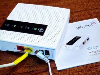 Ein Wlan-Router steht auf einem Holztisch, darunter eein Anleitungs-Büchlein. Der Router ist weis, quadratisch und hat ein schwarzes Bedienpanel mit mehreren LEDs auf der Obeseite, auf der Seite zwei Lanbuchsen rot und gelb, eine Reset-Öffnung, ein DC-Kabel steckt gewinkelt, ein ON/OFF-Schalter.