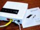 Ein Wlan-Router steht auf einem Holztisch, darunter eein Anleitungs-Büchlein. Der Router ist weis, quadratisch und hat ein schwarzes Bedienpanel mit mehreren LEDs auf der Obeseite, auf der Seite zwei Lanbuchsen rot und gelb, eine Reset-Öffnung, ein DC-Kabel steckt gewinkelt, ein ON/OFF-Schalter.