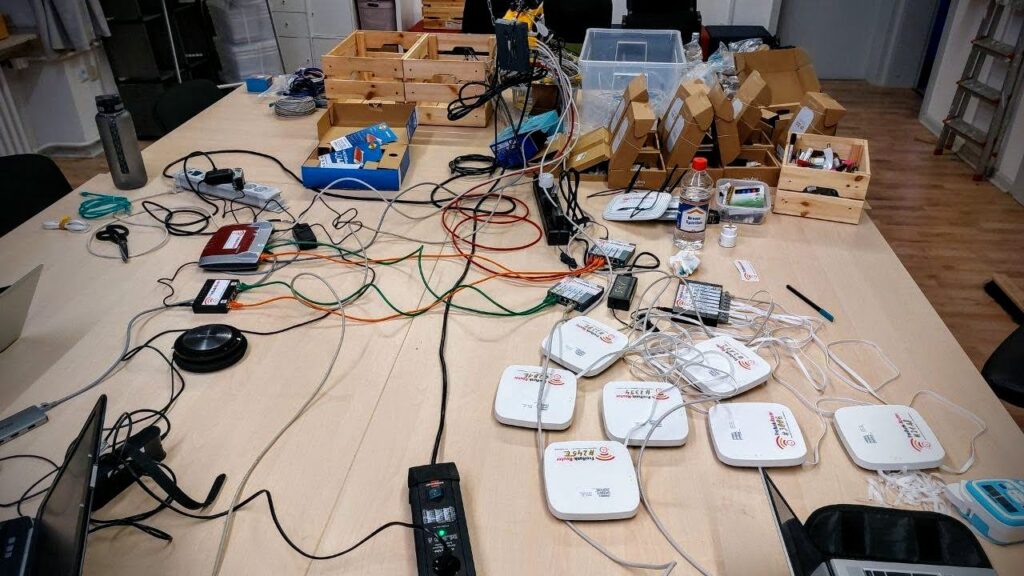 Foto eines großen Arbeitsgruppentisches. Auf dem Tisch zahlreiche Wlan-Router, Wlan-Accesspoints, Switches, Kabel, Laptop, Verteilersteckdosen. In Ikea-Holzkisten abgepacktes Installationsmaterial. Foto ist von vorangegangener Installationsparty.