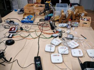 Foto eines großen Arbeitsgruppentisches. Auf dem Tisch zahlreiche Wlan-Router, Wlan-Accesspoints, Switches, Kabel, Laptop, Verteilersteckdosen. In Ikea-Holzkisten abgepacktes Installationsmaterial. Foto ist von vorangegangener Installationsparty.