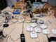 Foto eines großen Arbeitsgruppentisches. Auf dem Tisch zahlreiche Wlan-Router, Wlan-Accesspoints, Switches, Kabel, Laptop, Verteilersteckdosen. In Ikea-Holzkisten abgepacktes Installationsmaterial. Foto ist von vorangegangener Installationsparty.