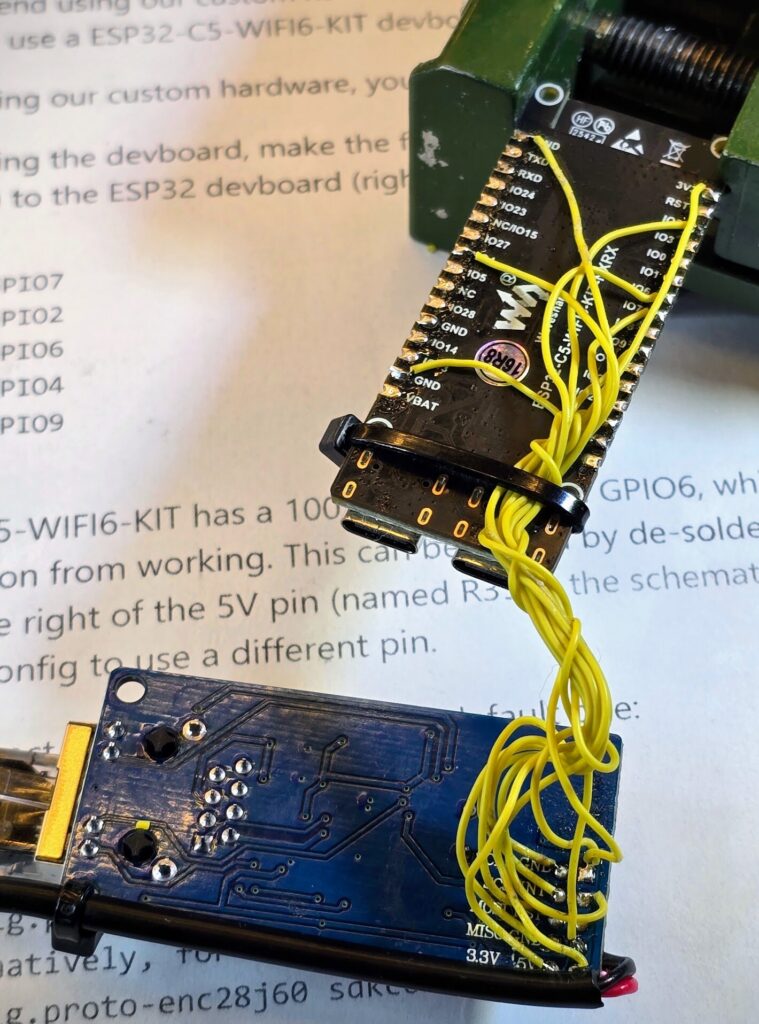 Zwei Platinen beim Verlöten: ein ESP32-C5-Board in einem kleinen Schraubstock, verbunden über viele gelbe Litzen mit einer Netzwerk-Platine. Darunter ein ausgedruckter Teil der Bauanleitung.