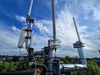 Antennenmast auf dem Dach des WIR-Hauses vor blauem Himmel: mehrere Rundstrahlantennen an Auslegern, links das weiße Gehäuse einer CPE210 mit dem Car2X-Empfänger, im Hintergrund Dächer und Bäume von Wülfrath