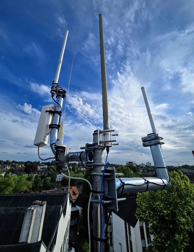 Antennenmast auf dem Dach des WIR-Hauses vor blauem Himmel: mehrere Rundstrahlantennen an Auslegern, links das weiße Gehäuse einer CPE210 mit dem Car2X-Empfänger, im Hintergrund Dächer und Bäume von Wülfrath